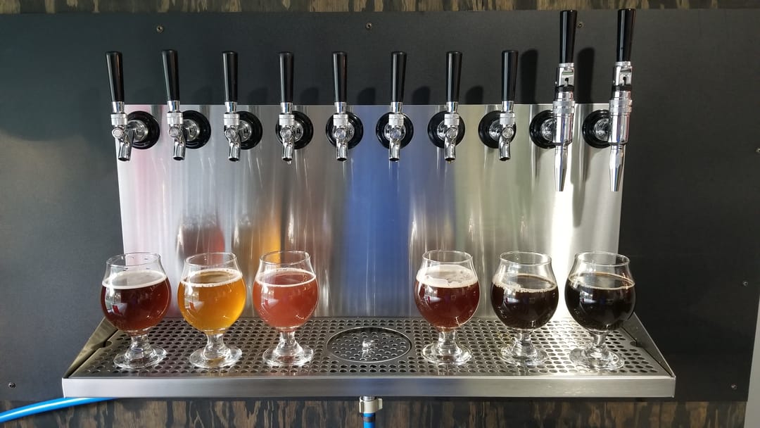 Six glasses of beer in a range of colors beneath the brewery taps